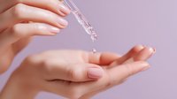 Female hand applying transparent serum from dropper pipette onto palm against soft purple background. Skincare routine demonstration.