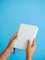 Hands holding blank white notebook with black bookmark against bright blue background for branding and mockup purposes.