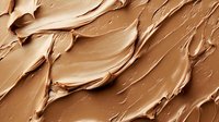 Creamy beige foundation texture with smooth swirls and strokes showing cosmetic product consistency, makeup background pattern with light and shadows.