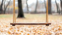 Empty wooden swing in autumn park with fallen yellow leaves on ground, blurred trees in background creating nostalgic mood and peaceful atmosphere.
