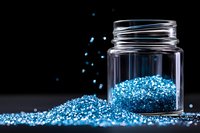 Shimmering blue glitter scattered around clear glass jar on dark background with magical sparkle effect, creating luxurious cosmetic or craft styling composition.