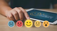 Customer feedback rating concept with colorful emoji faces from sad to happy on wooden desk next to tablet showing satisfaction survey interface.