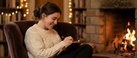 Young woman reading book in cozy armchair by fireplace with warm lighting creating peaceful evening atmosphere for relaxation and leisure activities.