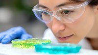 Laboratory scientist in protective glasses examining colorful petri dishes with bacterial cultures and growth media during microbiology research experiment.