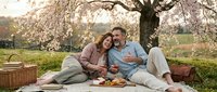A mature couple relaxes together on a blanket beneath a blooming tree during spring picnic, sharing food and quality time outdoors in a scenic park setting.