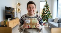 Happy middle-aged Caucasian man in festive sweater holding wrapped gift in decorated Christmas living room with tree and lights.