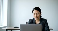 Young woman of East Asian descent, professional businesswoman wearing dark blazer, focused on laptop work at desk in modern office environment with natural window light.
