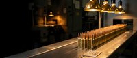Rows of golden brass rifle ammunition cartridges lined up on wooden surface in dimly lit workshop with warm pendant lighting creating dramatic shadows.