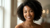 Happy young African American woman with curly hair smiling warmly in bright indoor setting. Professional headshot for business and lifestyle content.