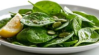 Fresh baby spinach salad with pumpkin seeds and lemon slice on white ceramic plate, close up view. Healthy green organic food with natural garnish.
