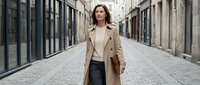 Young professional woman in beige trench coat walking confidently through modern urban street with contemporary architecture for business lifestyle content.