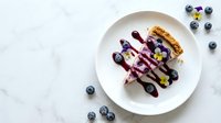 Blueberry cheesecake slice with fresh berries and sauce drizzle on white plate, overhead view on marble surface for dessert menu and food photography.
