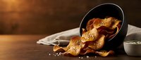Golden crispy potato chips spilling from black bowl on rustic wooden table with warm lighting and sea salt for snack food and cooking concepts.