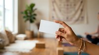 Woman with medium skin tone holding blank white business card in modern home office with natural light, plants, and neutral decor for branding mockups.