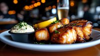 Grilled scallops with herb butter sauce and roasted vegetables on dark plate, fine dining restaurant setting with warm bokeh lighting and wine glass.