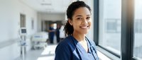 Young Hispanic female doctor smiling confidently in modern hospital corridor wearing navy scrubs and stethoscope for healthcare marketing materials.