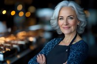 Confident mature Caucasian businesswoman with silver hair smiling in modern office environment with warm bokeh lighting background for corporate success.