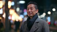 Mature Asian businessman in elegant coat and scarf walking through illuminated city street at night with warm bokeh lights creating atmospheric urban scene.