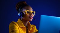Young African American woman customer service representative in yellow jacket and stylish glasses working at laptop with headset against blue background.