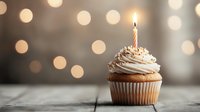 Vanilla cupcake with whipped cream frosting and lit birthday candle on rustic wooden surface against soft bokeh lights background, celebration mood.