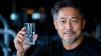Middle-aged Asian man with gray beard wearing black shirt holds up glass of water with ice cubes against dark background, confident expression and natural lighting.