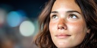 Young woman with freckles looking up thoughtfully against blurred bokeh background, natural beauty portrait with soft lighting and dreamy expression.