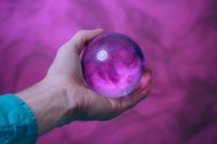 Hand holding transparent crystal ball with glowing purple smoke effect inside against mystical purple background, creating magical fortune telling atmosphere.