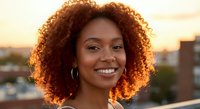 Young African American woman with curly hair smiling at golden hour sunset in urban setting with warm lighting and city skyline background.