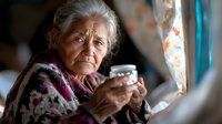 Elderly woman with gray hair holding cup by window, wrapped in colorful knitted shawl, contemplative expression showing life experience and solitude.