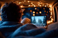 Cozy evening in car with laptop and fairy lights, watching movie under blanket. Back view of person relaxing during winter road trip or camping adventure.