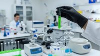 Scientist in black gloves holding green test tube in modern laboratory with equipment and researcher working in background for medical research.