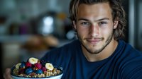 Young Caucasian man with stubble and wavy hair smiling while holding bowl of healthy breakfast with fresh berries, banana slices, and granola in dark moody lighting.