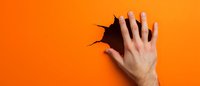 Female hand breaking through bright orange paper background creating dramatic torn hole effect, minimal composition for advertising and design projects.