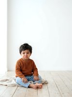 Young boy with dark hair and medium skin tone, wearing rust-orange ribbed sweater and light denim jeans, sitting cross-legged on wooden floor against white wall. Child appears upset or pouting. Isolated on white background, suitable for parenting, childcare, emotions, and family-related design projects.