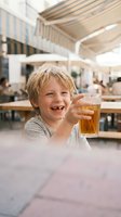 Happy blonde boy holding glass of apple juice at outdoor restaurant terrace during summer vacation with family dining experience.