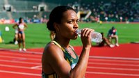 Female athlete of South Asian descent drinking water during track and field competition at stadium, refreshing during athletic event for sports hydration and endurance training.