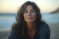Confident middle-aged woman with natural wavy brown hair smiling gently at sunset on beach, wearing casual dark sweater. Soft golden hour lighting creates warm atmospheric portrait.