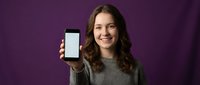 Happy young Caucasian woman with curly brown hair holding smartphone with blank white screen against purple background for mockup and app presentation.