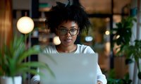 Young African American businesswoman with natural curly hair and glasses working on laptop in modern coworking space with ambient lighting and plants.