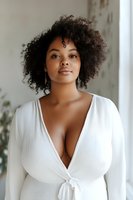 Young African American woman with natural curly hair wearing white wrap dress poses confidently in soft window light, expressing self-acceptance and modern beauty.