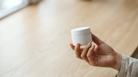 Person with medium skin tone holding white cosmetic jar with lid against wooden surface background for skincare product marketing.