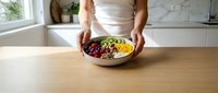 Woman in white top holding a colorful fruit and nut bowl on a wooden kitchen table, healthy eating concept for wellness blogs.