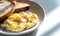 Creamy scrambled eggs with black pepper served on white ceramic plate alongside toasted bread slices, bathed in natural morning light. Fresh breakfast setting.