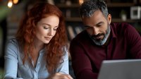 Woman with red curly hair and man with gray beard working together on laptop in office setting. Diverse colleagues collaborating on project.