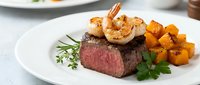 Surf and turf surf and turf dish with seared shrimp on top of medium-rare beef steak, served with roasted sweet potato cubes and fresh herbs on white plate.