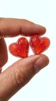 Close-up of hand holding two red heart-shaped gummy candies with sparkling sugar coating on white background. Isolated for design use.