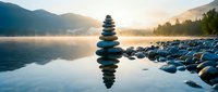 Zen stone stack balanced on calm lake water with mountain sunrise reflection creating peaceful meditation atmosphere for wellness and mindfulness concepts.