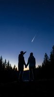 Couple holding hands watching meteor streak across night sky above forest at dusk, romantic stargazing moment for astronomy and relationship themes.