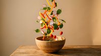 Fresh vegetables falling into wooden bowl on rustic table with warm lighting for healthy cooking and meal preparation concepts.