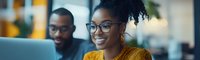 Young African American businesswoman wearing glasses and yellow blouse smiling during office meeting with colleague, panoramic corporate workspace background.